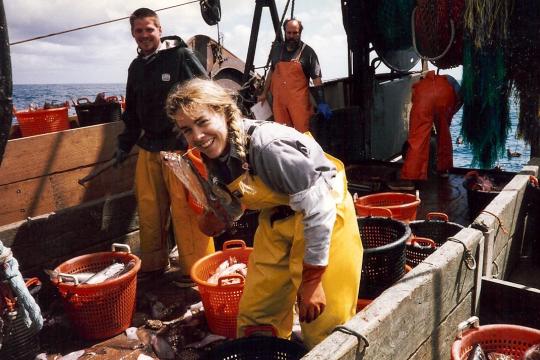 A woman poses holding a fish while aboard a fishing vessel with fisherman and their catch separated into baskets in the background. 