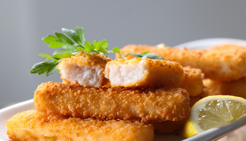Fried fish sticks stacked on a white plate. One fish stick is split in half at the stop of the stack to show the white fish inside. The plate is garnished with a lemon wedge and parsley