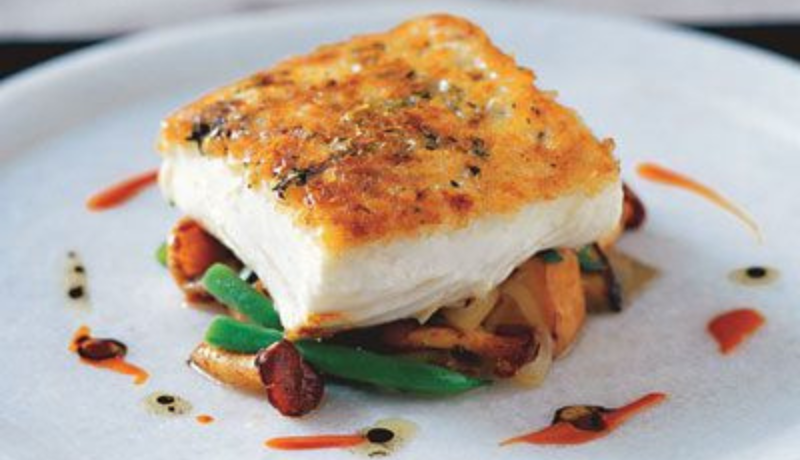 A white plate with a white fish filet on a bed of orange, green, and white sauteed vegetables. The fish filet has a brown crusted top. Around the food is a drizzle of orange and white oils with black dots.